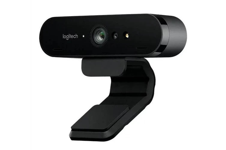 A black Home Office Space NZ Logitech BRIO Ultra HD Webcam with a rectangular design is mounted on an adjustable stand. The camera lens, which captures 4K Ultra HD video, is centered and flanked by two smaller sensors and noise-canceling microphones. The Logitech logo is visible on the left side of the device. The background is white.