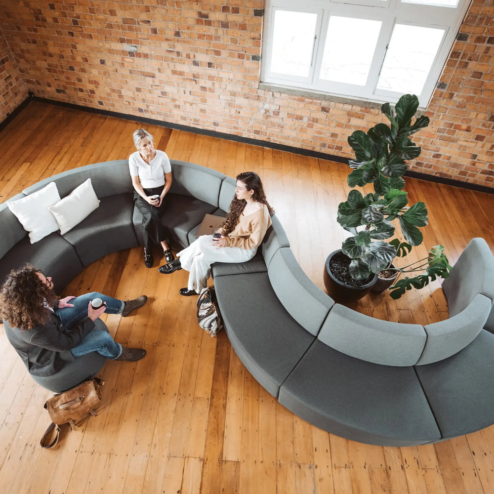 Three people are sitting and conversing on a curved sectional sofa from the Konfurb Arco Series by Home Office Space NZ in a spacious room with brick walls and wooden floors. A large potted plant is placed nearby. Two of them are sitting with laptops, while one holds a coffee cup, enjoying the exceptional design of this modular seating system, perfect for evolving work environments.