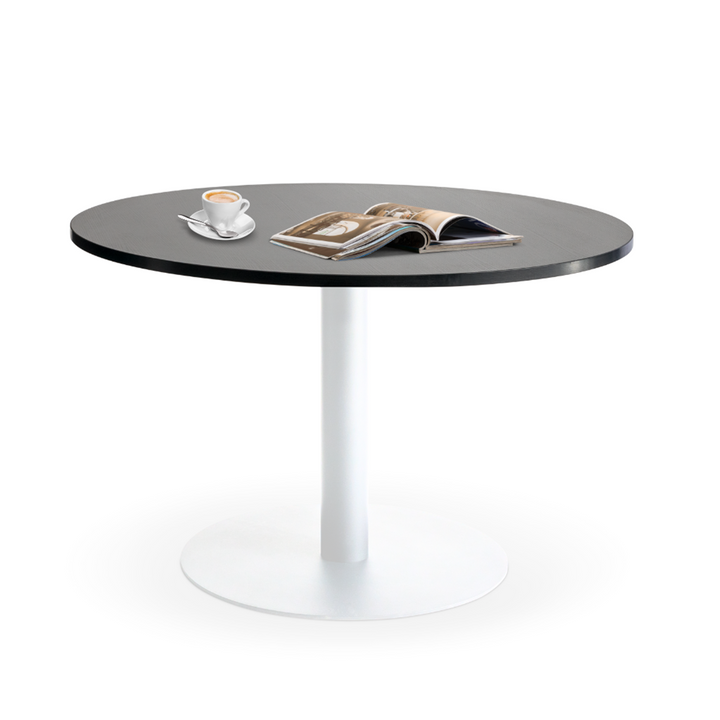 Introducing the Lunar 1200 Meeting Table by Home Office Space NZ, featuring a robust black melamine round top and a sleek white pedestal base. On the table, you'll find a small cup of cappuccino on a saucer and an open magazine with gently curved pages. The minimalist white backdrop makes this table an ideal choice for your home office space.