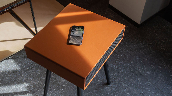 A smartphone sits on the MySound Blues Bluetooth Speaker Side-Table by Home Office Space NZ, which features angled black legs on a speckled gray floor. Sunlight highlights its sleek design and integrated sound system, while it seamlessly offers wireless charging for devices.