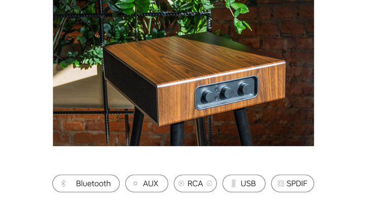 The MySound Blues Bluetooth Speaker Side-Table by Home Office Space NZ sits on a table, serving as an integrated sound system with three front control knobs, and icons for Bluetooth, AUX, RCA, USB, and SPDIF connections. The scene features a brick wall background and lush plants.